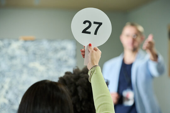Focus on hand of young female participant of auction raising paddle with number twenty seven in front of camera