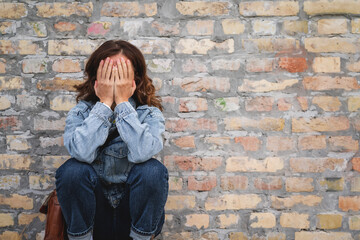 Burnt-out urban woman with her face covered by her hands sits next to a brick wall.
