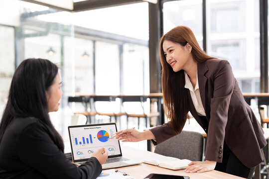Two happy asian businesswomen coworking with a laptop in a desktop at office, discussing and consulting business plan with graph chart.