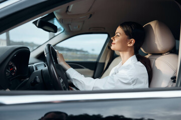 A woman is seated in the driver's seat of a car, hand on the steering wheel, gazing thoughtfully to...
