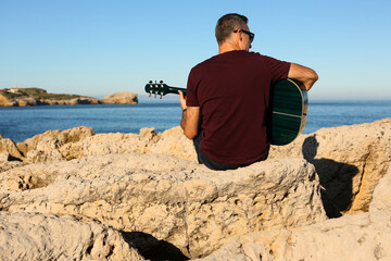A man in a dark t-shirt playing acoustic guitar on a rocky area by the sea