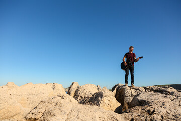 A man in a dark t-shirt playing acoustic guitar on a rocky area by the sea
