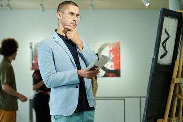 Fototapeta premium Young pensive male visitor of gallery in blue elegant suit holding brochure and looking at artwork at exhibition