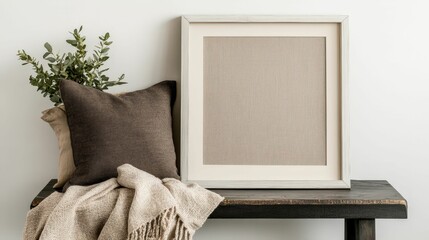 A cozy interior scene featuring a framed textured artwork, a decorative pillow, and a soft throw, all arranged on a wooden table with greenery accents.