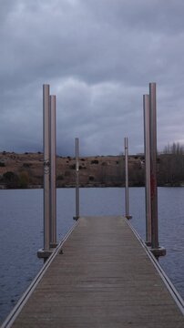 tranquil scene at Fuentes Claras in Avila, featuring a wooden dock extending into a calm lake. The overcast sky and barren hills in the background create a peaceful and reflective atmosphere