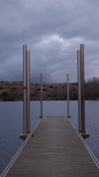 tranquil scene at Fuentes Claras in Avila, featuring a wooden dock extending into a calm lake. The overcast sky and barren hills in the background create a peaceful and reflective atmosphere