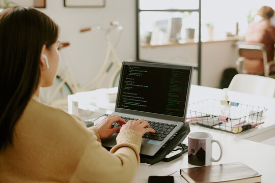 Back view of unrecognizable female office worker sitting at table and writing code on laptop with cooling pad