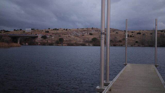tranquil scene at Fuentes Claras in Avila, featuring a wooden dock extending into a calm lake. The overcast sky and barren hills in the background create a peaceful and reflective atmosphere