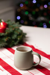 A cup with hot drink on white table with advent wreath and red candles, blurry background with Christmas tree lights