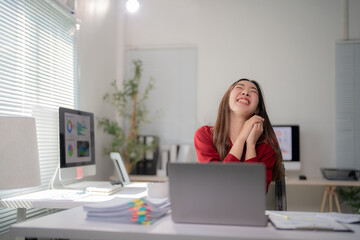 Young businesswoman is sitting at her desk in a modern office, laughing with joy and clasping her...