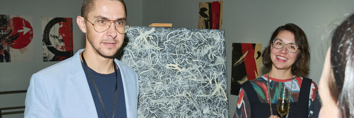 Header of young male worker of modern art gallery talking to public while making presentation of new painting