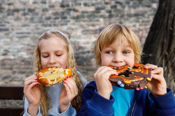 Portrait two happy funny playful blond children enjoy eating pretzels chocolate toppings at city street outdoor park. Scenic fun moment capturing childhood joy snacking on colorful treats