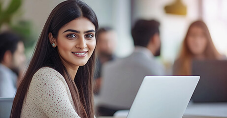 Fototapeta premium A close-up of a smiling woman with long dark hair, seated at a desk with a laptop. Professional and collaborative office environment in the background