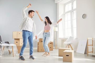 Young couple celebrating moving day smiling and dancing in new apartment full of cardboard boxes....