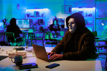 Medium close up of hacker wearing black hoodie and white mask hiding his face programming in dark...