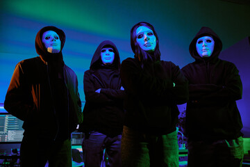 Low angle portrait of hacker group wearing black hoodie and white mask to hide their faces standing...