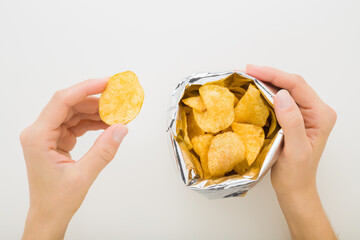 Young adult woman hands holding yellow potato chips and opened foil bag on light gray table background. Closeup. Point of view shot. Top down view.