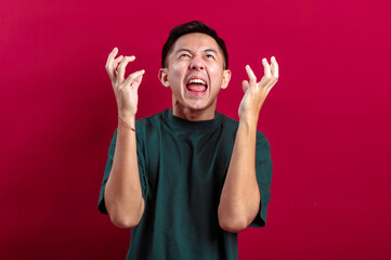 Fototapeta premium Asian man in a green T-shirt clenching his fists and showing an angry, frustrated expression against a vibrant red background, expressing intense emotion or irritation in a dramatic and relatable way
