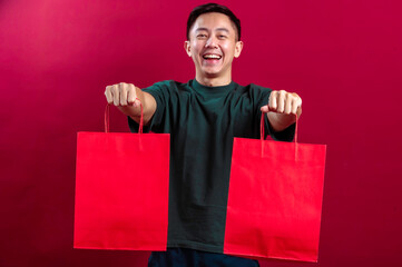 Asian man in a green T-shirt smiling cheerfully, holding vibrant red shopping bags in his hand, posing confidently with a hand on his hip, against a bold red background.