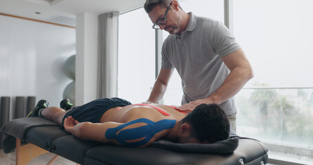 Physiotherapist, person and tape on bed for injury with recovery process, back pain and help at clinic. People, biokinetics and treatment for rehabilitation with muscle, spine and service at hospital