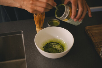 Woman adding matcha powder into bowl. Girl make atcha tea latte in the kitchen. Close up. Image matcha tea powder pouring in bowl. Japanese green tea concept.