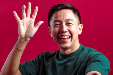 Naklejka premium Asian man in a green T-shirt smiling while holding a smartphone in one hand, raised for a selfie or video call, and waving with the other hand, against a vibrant red background