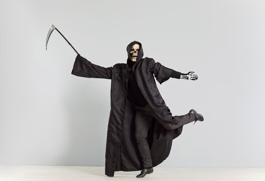 Grim reaper, holding a scythe and wearing a skeleton halloween mask, dancing on a grey background. Horror themed image captures the essence of death and the devil in a macabre, eerie dance.