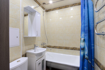 Bathroom in the apartment. Interior of a combined bathroom. White bathtub and sink with a tap. On the wall is a hanging cabinet with a mirror. The walls are decorated with tiles.