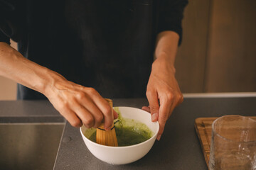 Woman cook matcha tea latte at home with bowl and bamboo whist. Happy girl with high bun mix matcha powder in bowl in the kitchen.
