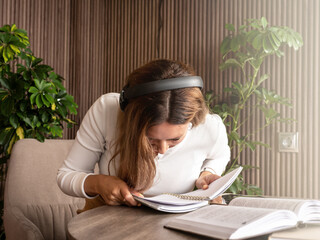 Woman in headphones sitting at table, bending closely over a notebook while reading or studying,...