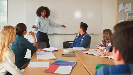 Female Tutor At Whiteboard Teaching Class Of High School College Or University Students Around Table
