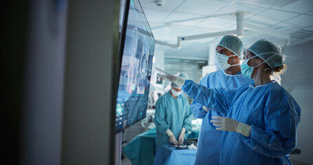 Portrait of a Young Female and Male Surgeons Reviewing Patient's Scans on a Large Screen in a Dimly...