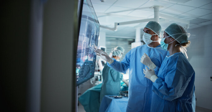 Caucasian Male and Female Surgeons Analyze Scans on a Digital Monitor, Planning Surgical Strategy in a High-tech Operating Room. Colleagues Working Together, Preparing for Surgery