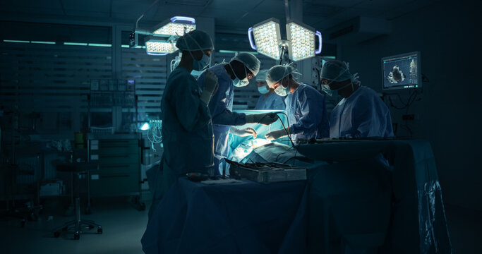 Dark Operating Room with a Diverse Team, Including a Black Male Assistant and a White Female Surgeon, Perform a Precise Surgical Procedure as Teamwork and Under Surgical Light