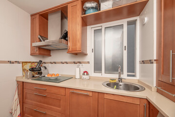 L-shaped fitted kitchen with cream stone effect worktop, sliding window with sink below, stainless steel appliances and cherry wood cabinets.