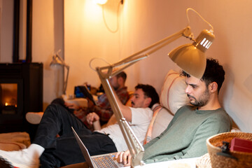 three men sitting on a large sofa working on a computer, looking at the mobile phone, disconnected from each other, in a warm and comfortable home-working environment.