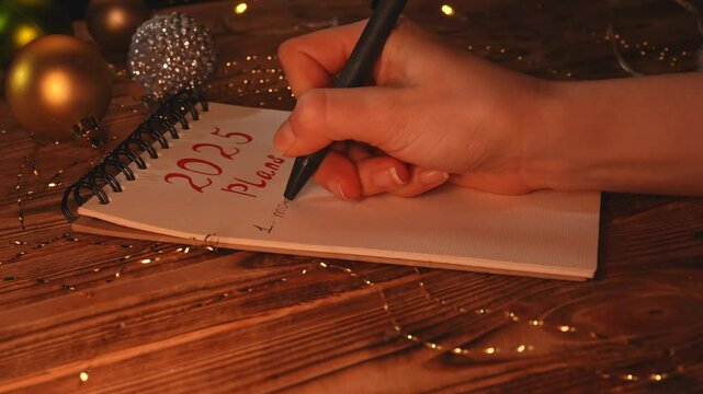 Close up shot of christmas decorated wooden table, woman writing with pen in spiral notebook plans for new 2025 year, numbering list with love, happiness and money.