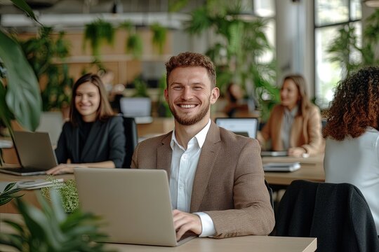 Smiling young man working on laptop in modern office filled with greenery and fellow coworkers