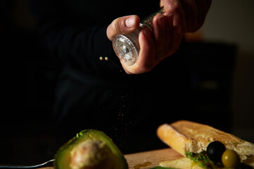 Hand sprinkling salt on fresh avocado near bread and olives
