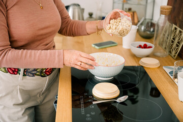 Elderly woman pouring oat flakes into bowl for breakfast in kitchen