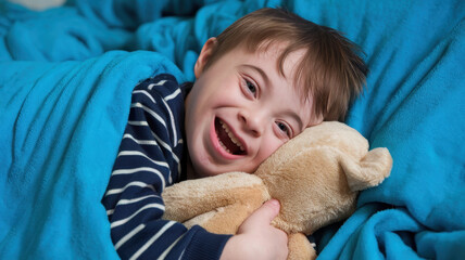 A joyful young boy with Down syndrome snuggles a teddy bear while wrapped in a cozy blue blanket. His cheerful smile fills the room with warmth.