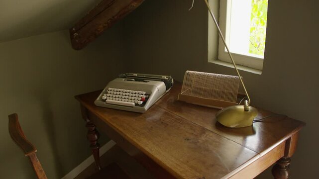 Cozy corner with a wooden desk featuring vintage typewriter, brass desk lamp, and wire organizer. Small window with greenery outside adds natural light, slanted ceiling creates a quaint atmosphere