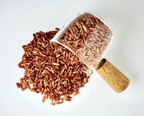 Pile of uncooked raw brown red rice or beras merah food ingredients spilled from small glassware cup pot container isolated on horizontal white background from top angle view.