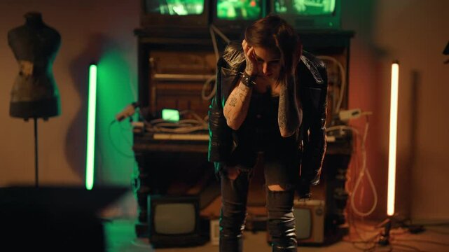 Lonely depressed woman standing alone in strange studio or basement, portrait. Alternative style, rock and punk aesthetics, young adult woman with tattoos on body holding head by hands, feeling bad