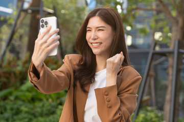 A joyful Asian businesswoman engages with her phone celebrating success in remote work. Smiling...