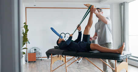 Physical therapy, men and band for stretching legs at clinic, helping and recovery on table. People, flexibility training and chiropractor for rehabilitation treatment, support and healing service