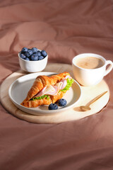 Fresh croissant sandwich with ham, cheese and lettuce, with cup of coffee and blueberries on tray. Breakfast in bed. Vertical orientation.