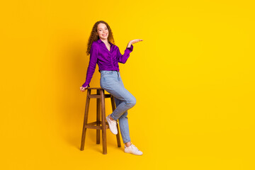 Fototapeta premium Full size photo of attractive young woman sit chair hold empty space dressed stylish violet clothes isolated on yellow color background