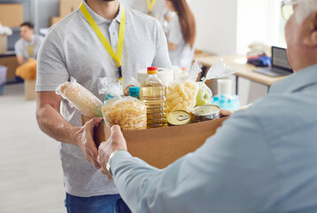 Volunteer working at charity center giving free food donations in cardboard box for mature man in charitable foundation. Humanitarian aid, volunteering and social help for poor concept.