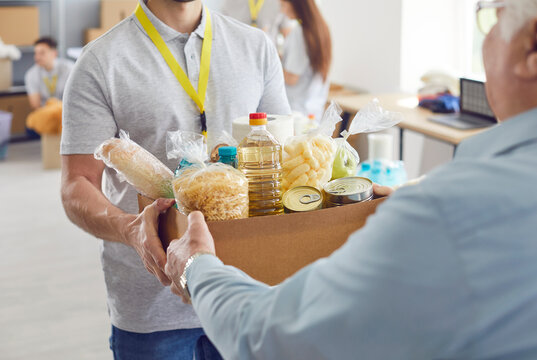 Volunteer working at charity center giving free food donations in cardboard box for mature man in charitable foundation. Humanitarian aid, volunteering and social help for poor concept.